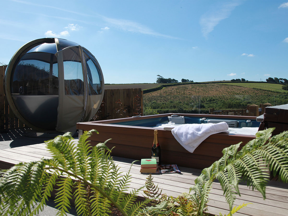 An outdoor hot tub and modern glass pod in a serene garden on a sunny day