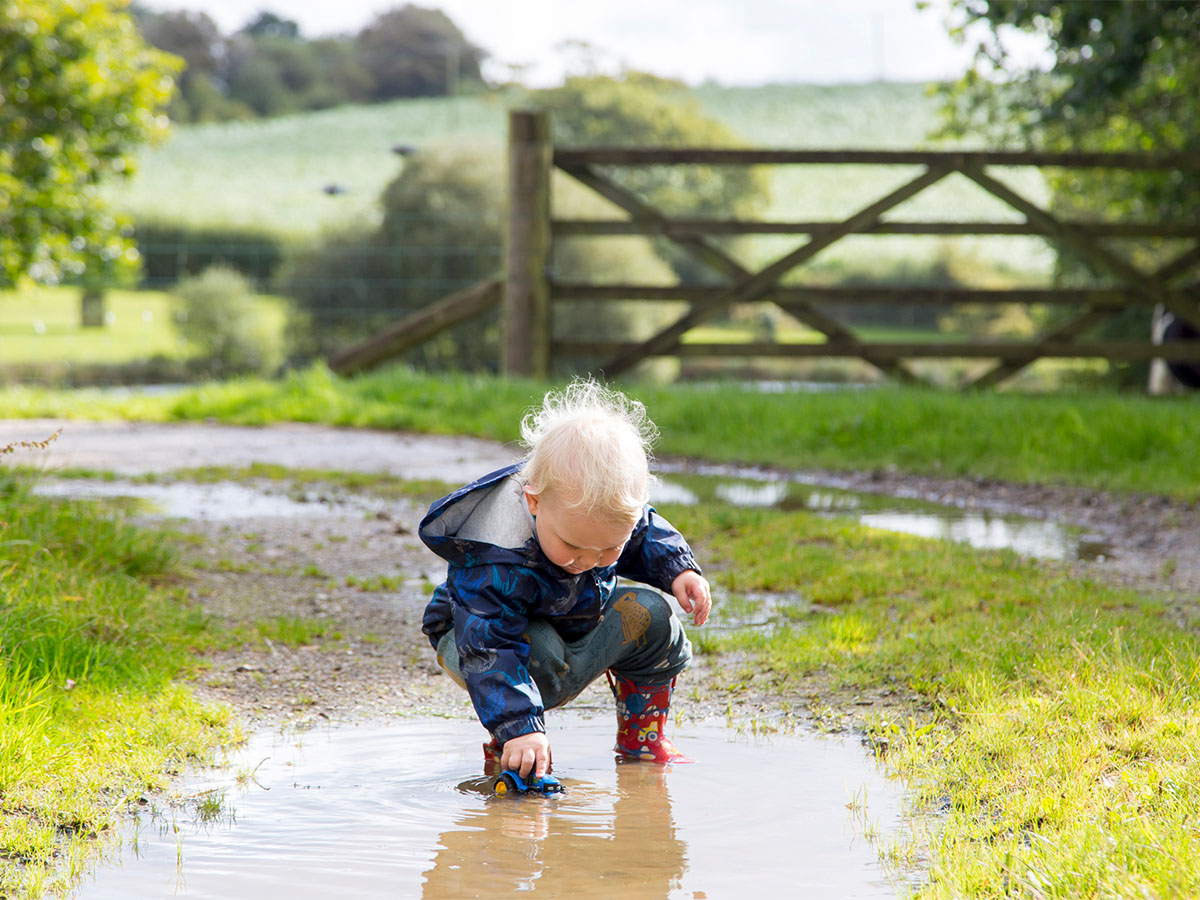 A little boy in a puddle suit splashes in a muddy puddle in front of a farm gate
