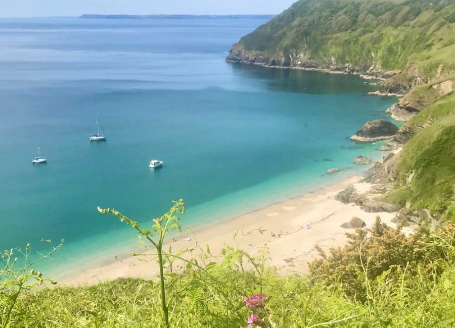 Lantic Bay in Cornwall. It's a sunny day and there are three boats on the water as well as people on the beach.