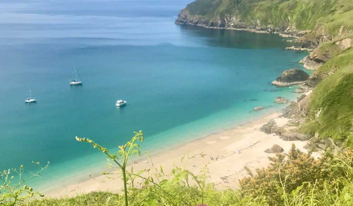 Lantic Bay in Cornwall. It's a sunny day and there are three boats on the water as well as people on the beach.