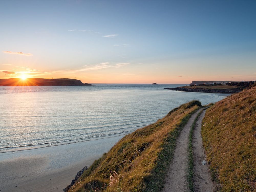 A stretch of the South West Coast Path heading towards Daymer Bay with the sun setting in the distance.