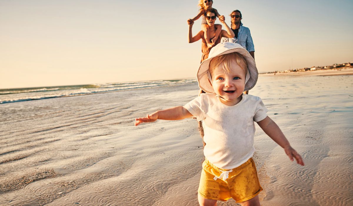 Young child smiling with a sun hat on and running towards the camera on a beach with family behind him at sunset